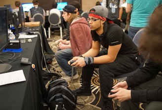 Several people are sitting in a room with gaming consoles and monitors, focusing intently on their screens. The setting appears to be a casual gaming event, with participants holding controllers while playing video games. The room has a casual atmosphere with chairs and tables set up for gaming.