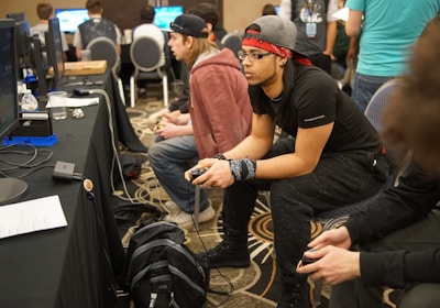 Several people are sitting in a room with gaming consoles and monitors, focusing intently on their screens. The setting appears to be a casual gaming event, with participants holding controllers while playing video games. The room has a casual atmosphere with chairs and tables set up for gaming.