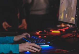 A person is engaged in a video game at a computer setup, using a keyboard and mouse. The scene is dimly lit, with the glow from the monitor reflecting off the hands of the player. Several other people are present in the background, indicating a social or competitive gaming environment.