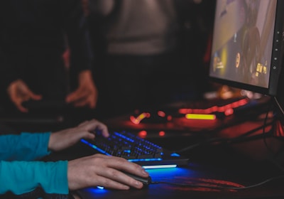 A person is engaged in a video game at a computer setup, using a keyboard and mouse. The scene is dimly lit, with the glow from the monitor reflecting off the hands of the player. Several other people are present in the background, indicating a social or competitive gaming environment.