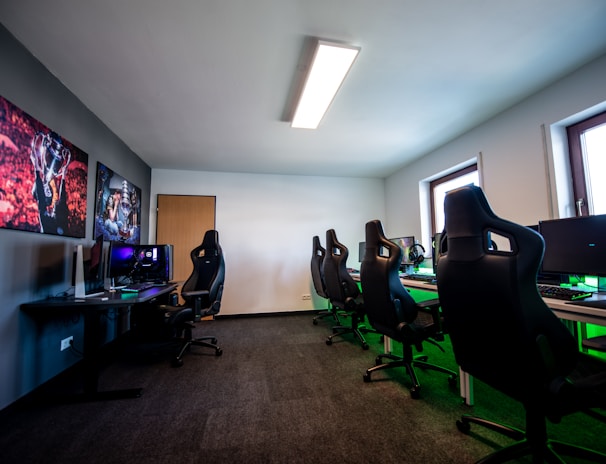 A well-lit gaming room features multiple gaming stations with high-backed chairs and monitors. The room has a minimalist setup with green and black color tones. Large posters adorn the walls, and the room appears clean and organized.