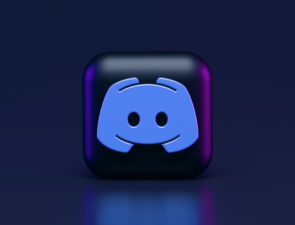 A 3D-style block with a blue logo in the center, set against a dark background. The logo resembles a game controller with two rounded edges and two circles on the front. The block has a subtle gradient, transitioning from dark to purple on its sides.