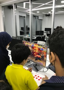 A group of children is gathered around a desktop computer, engaged in playing a colorful, action-packed arcade game displayed on the monitor. The setting appears to be a modern, well-lit office or gaming space with large glass windows, showing multiple computer workstations in the background.