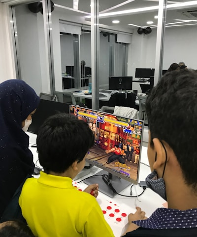 A group of children is gathered around a desktop computer, engaged in playing a colorful, action-packed arcade game displayed on the monitor. The setting appears to be a modern, well-lit office or gaming space with large glass windows, showing multiple computer workstations in the background.