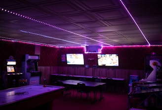 A dimly lit room with vibrant purple LED strip lights on the ceiling, featuring multiple gaming stations. There are two large screens showing video games, along with various arcade machines, including a Nintendo arcade cabinet. An air hockey table is positioned in the foreground.