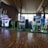A vibrant arcade setting with several gaming machines lined up, including dance and rhythm game setups. People are engaged with the games, absorbed in their activities. The flooring is wooden, and the ceiling has a geometric design. Natural light filters through large windows, creating a lively atmosphere.